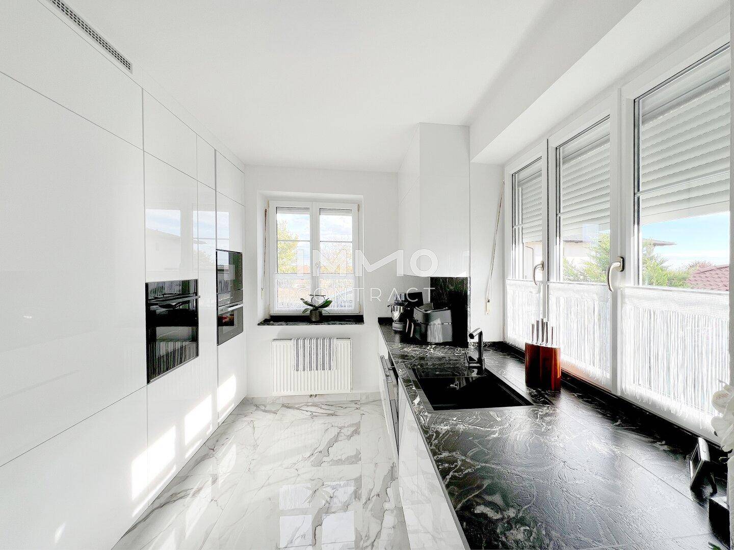 Moderne weiße Küche mit Hochglanzfronten, dunkler Arbeitsplatte und viel Tageslicht.