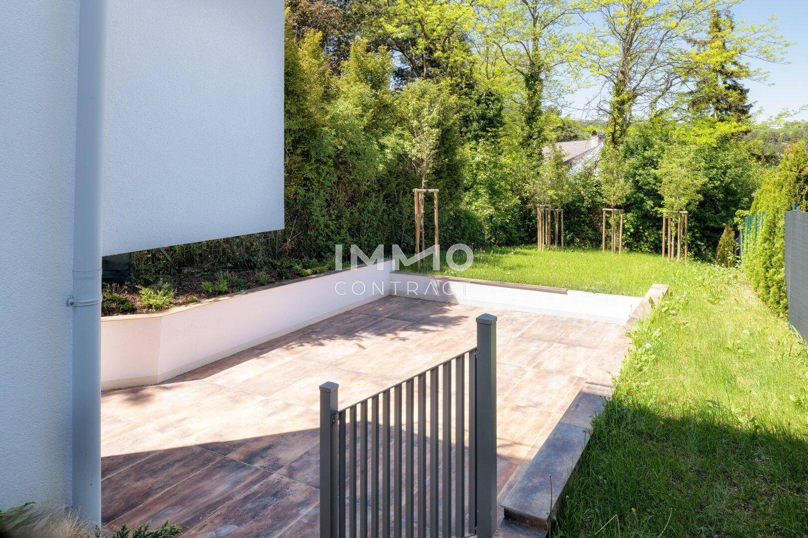 Sonniger Terrassenbereich mit Fliesenboden und angrenzender Rasenfläche, umgeben von Grün.