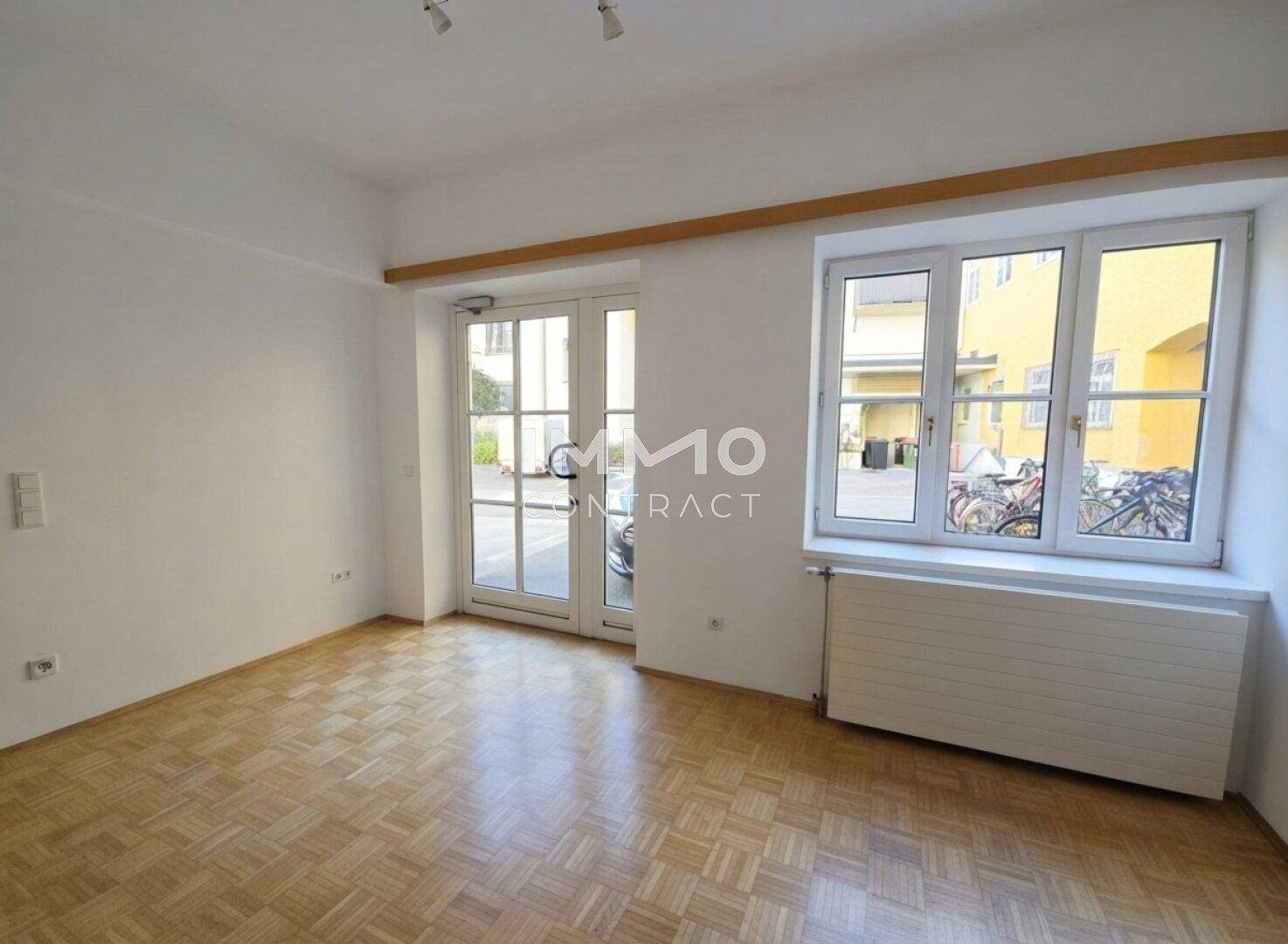Heller Raum mit Parkettboden, großen Fenstern und einer Glastür, die nach draußen führt.