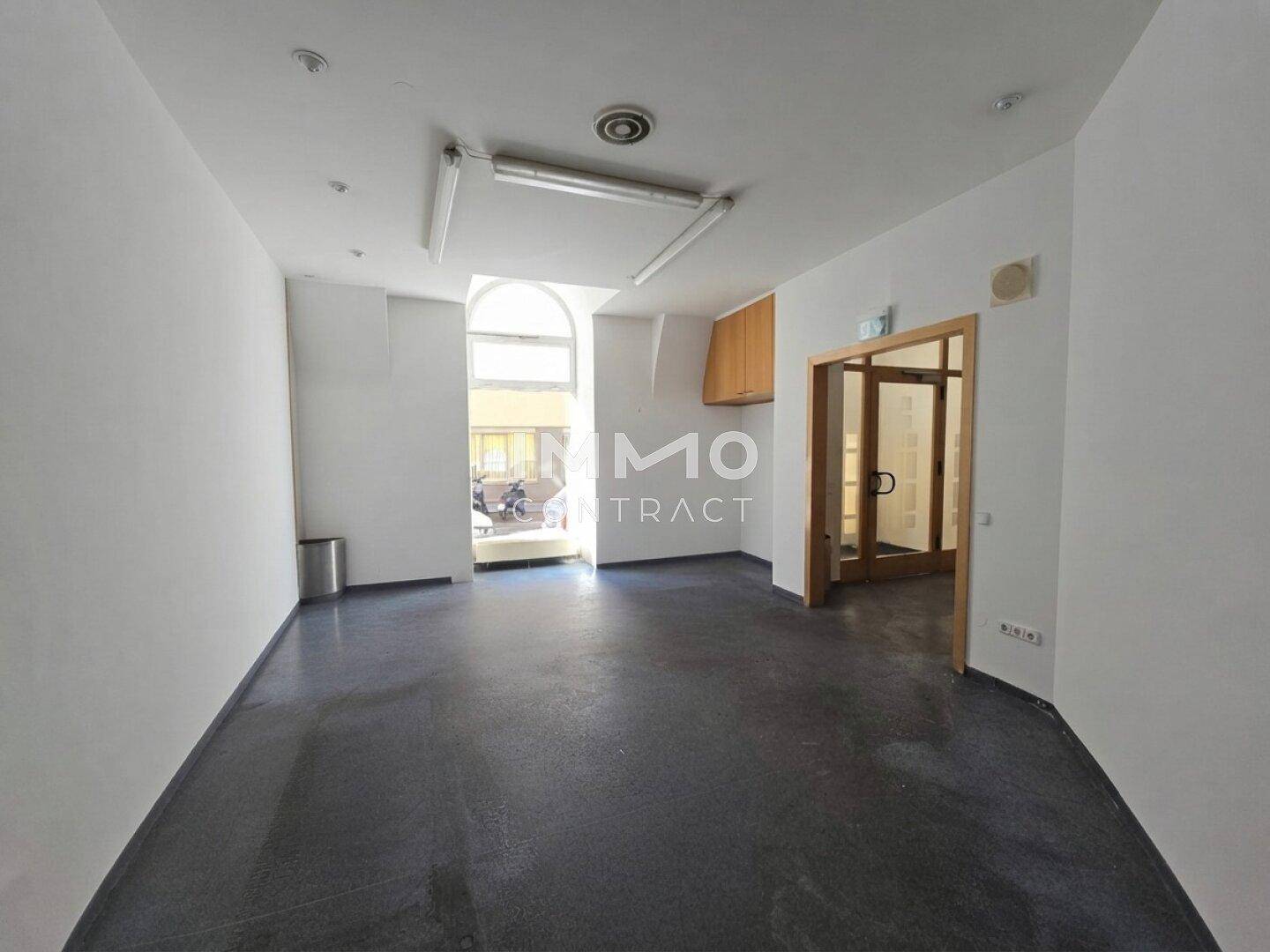 Heller Gewerberaum mit dunklem Boden, gewölbtem Fenster und kleiner Küchenzeile, bietet viel Tageslicht.