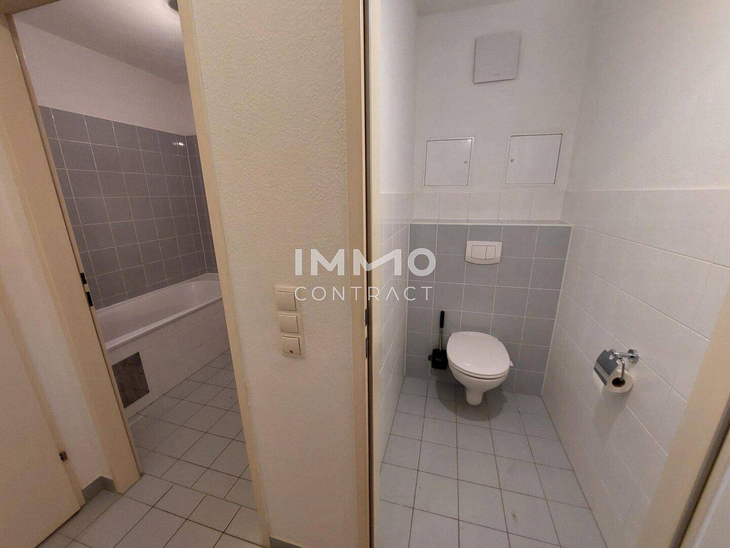 Getrennte Toilette mit hellgrauen Fliesen und Blick auf das angrenzende Badezimmer mit Badewanne.