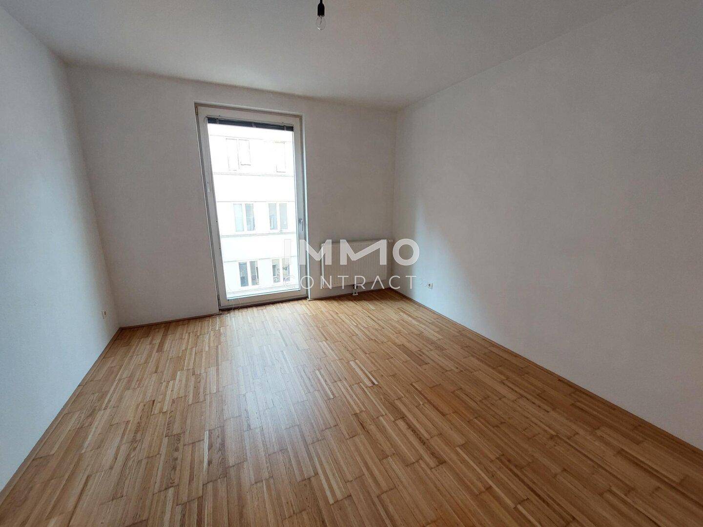 Leeres Schlafzimmer mit Parkettboden und einem großen Fenster, das viel Licht spendet.