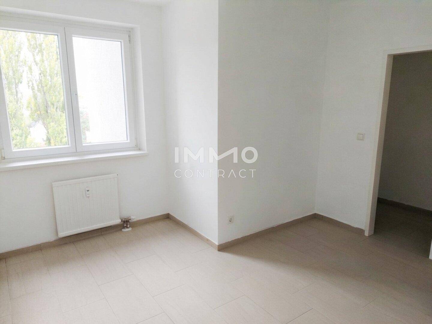 Leeres Zimmer mit hellem Fliesenboden, großem Fenster und Heizkörper.