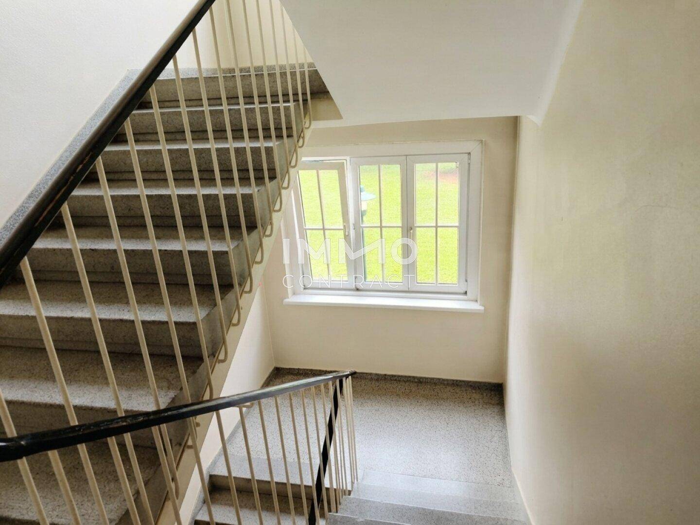 Treppenhaus mit Steinstufen und Metallgeländer, hell durch ein Fenster am Zwischenpodest.