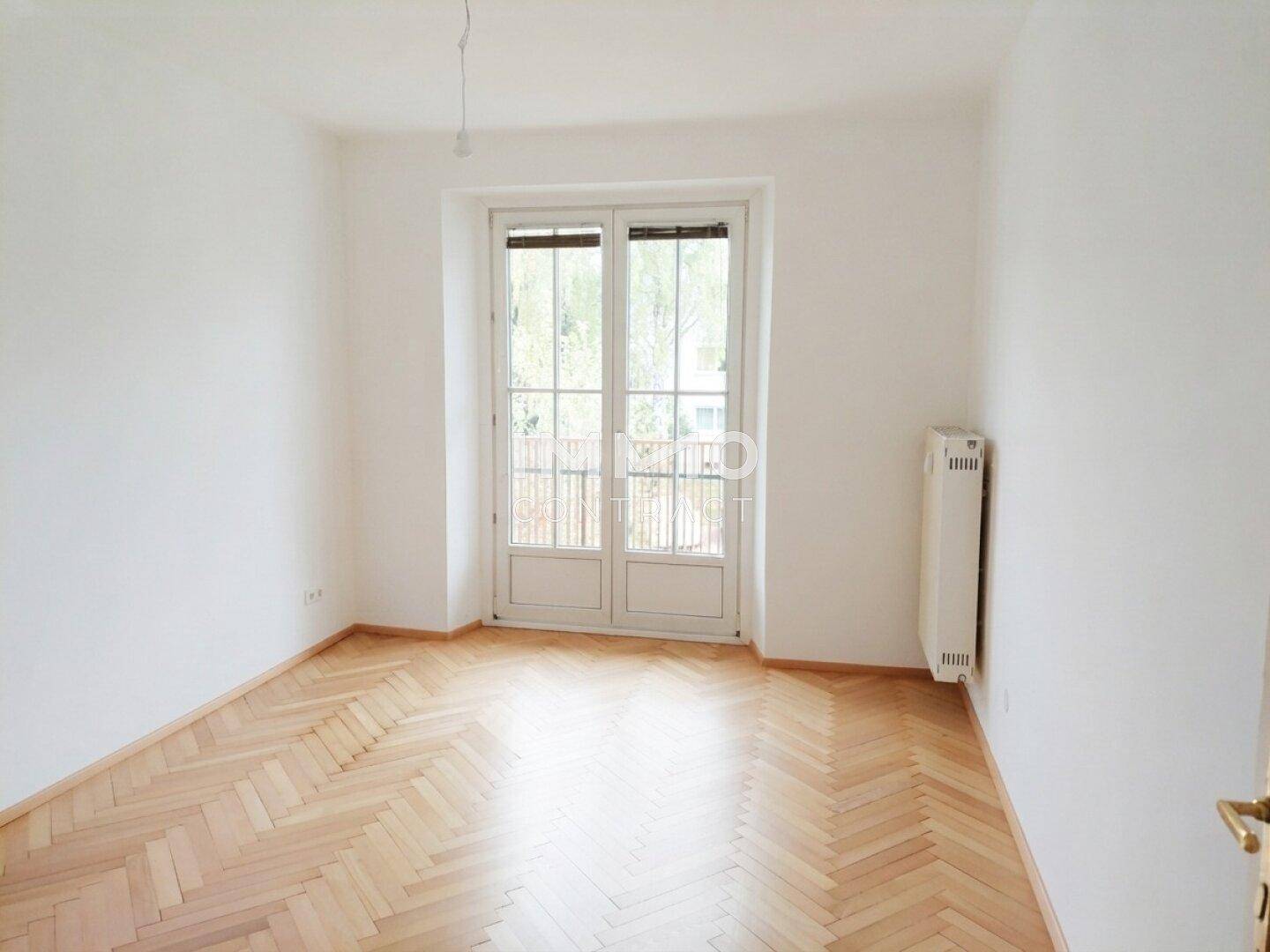 Leeres Zimmer mit Fischgrätparkett und Fenstertür, die zu einem französischen Balkon führt.