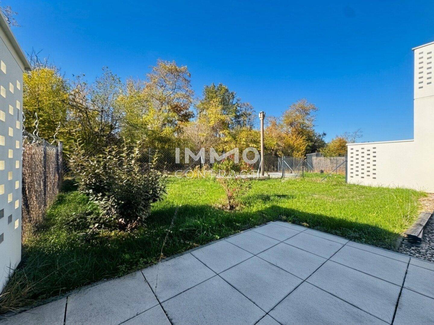 Sonniger Terrassenbereich mit Pflastersteinen und angrenzendem Rasen, umgeben von Bäumen.