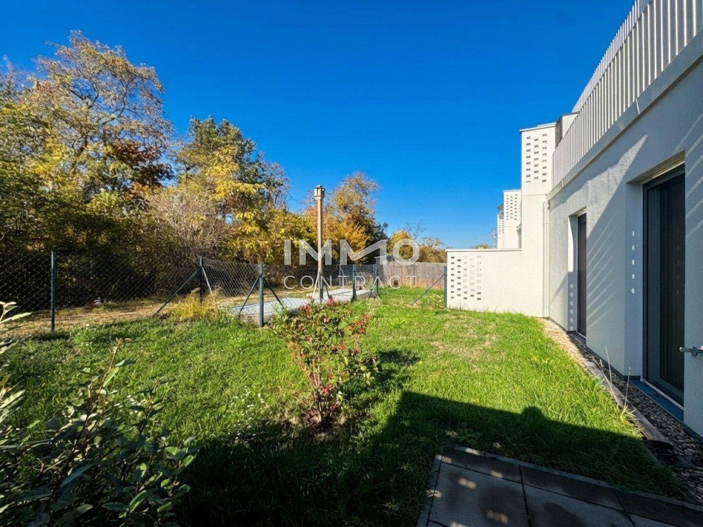 Grüner Gartenbereich neben dem Gebäude mit Rasenfläche und Bäumen im Hintergrund unter blauem Himmel.