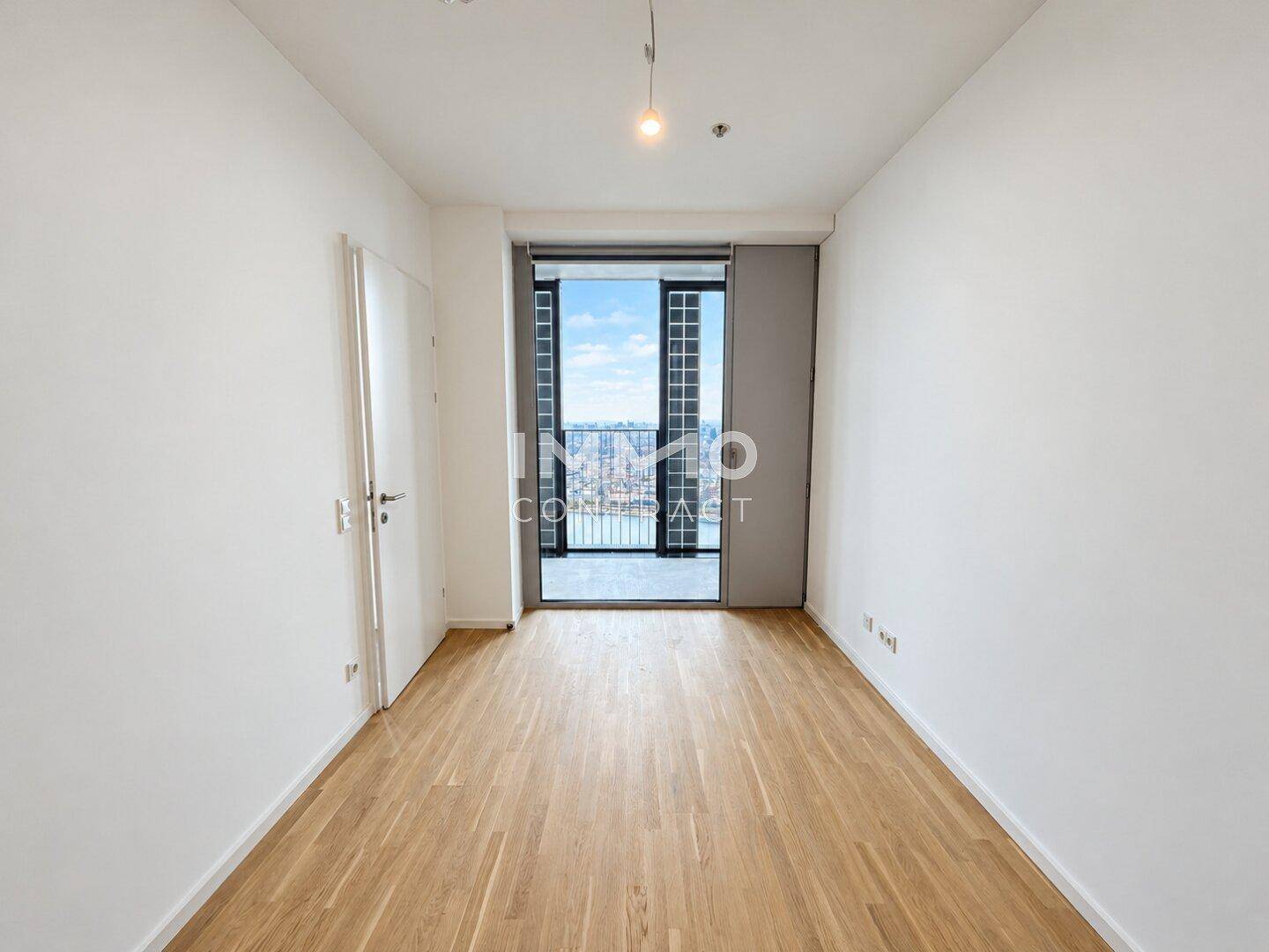 Helles Zimmer mit Parkettboden und großem Fenster, das einen weiten Blick über die Stadt ermöglicht.