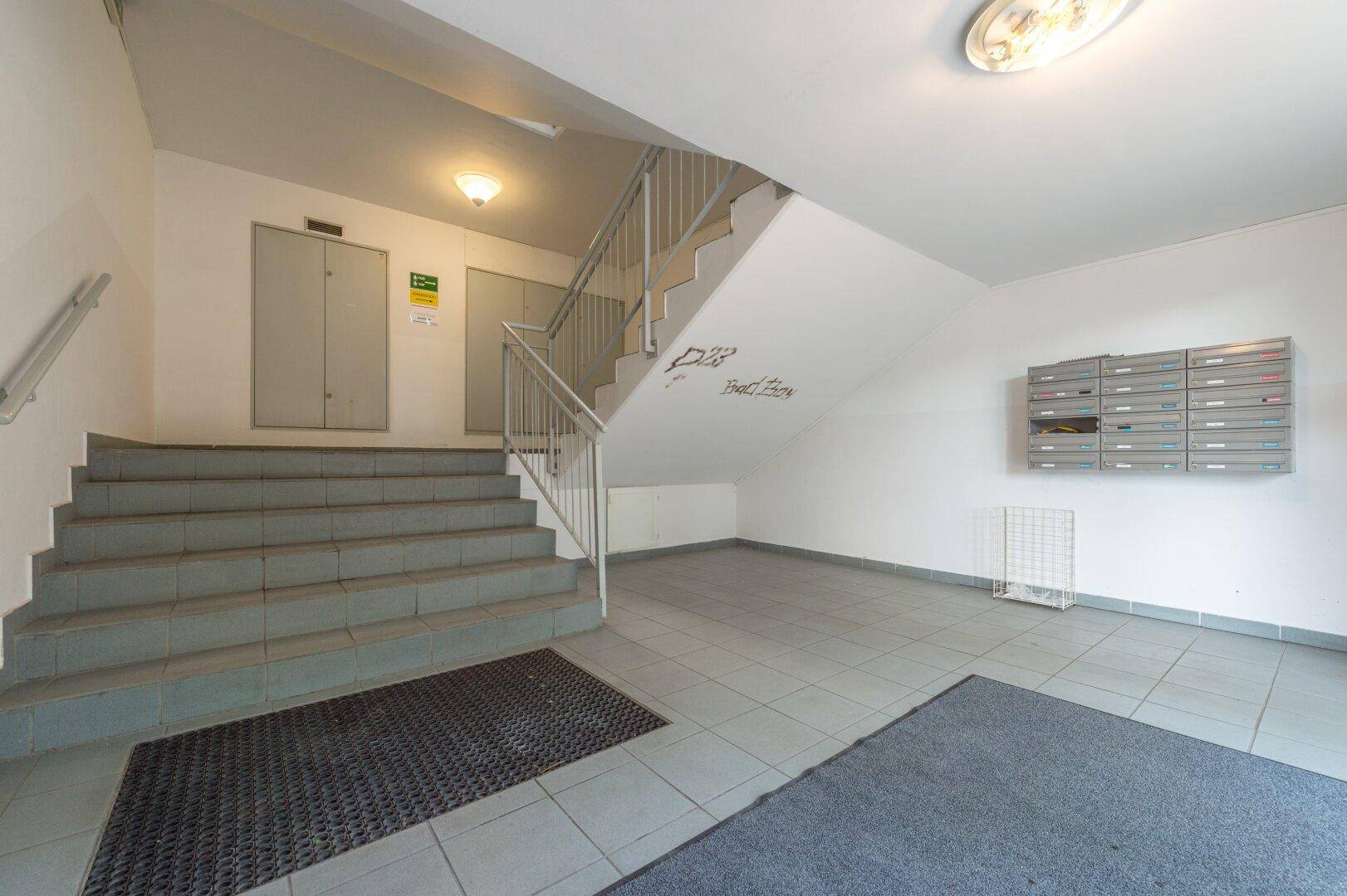 Treppenhaus mit grauen Fliesen, Betontreppen und einem Metallgeländer, inklusive Briefkästen.