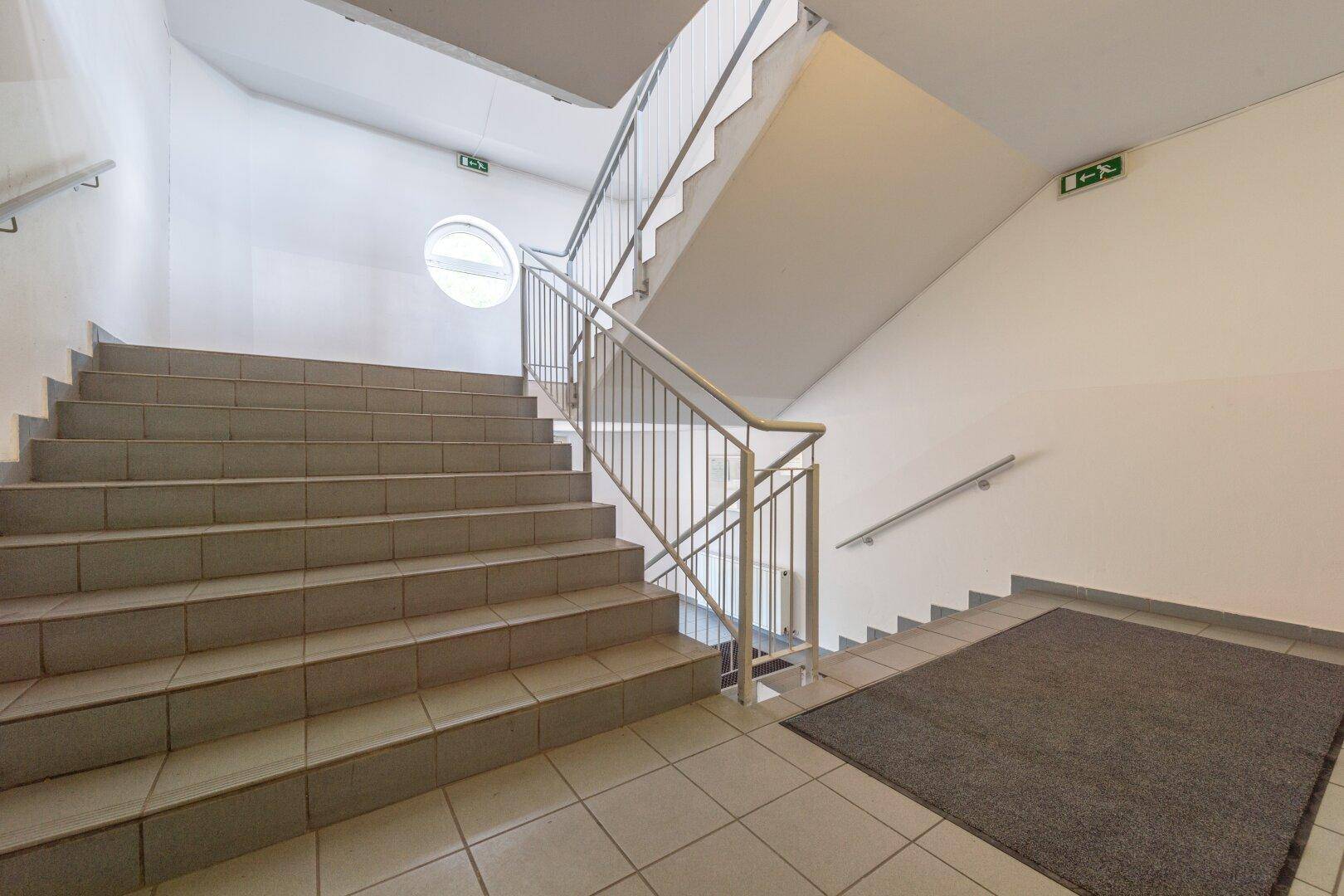 Helles Treppenhaus mit Fliesenboden, Betontreppen und einem runden Fenster für natürliches Licht.