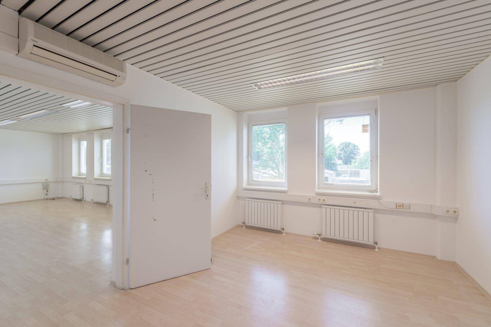 Heller Büroraum mit Klimaanlage, Holzoptikboden und Fenstern mit Blick auf die Umgebung.