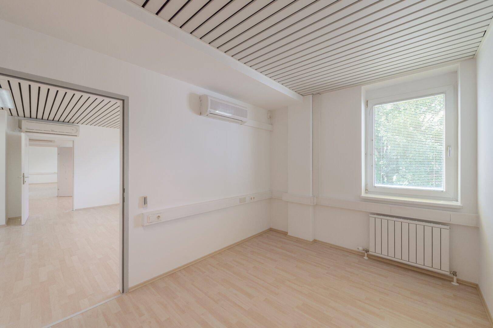Sauberer Büroraum mit Klimaanlage, Jalousien am Fenster und hellem Laminatboden.