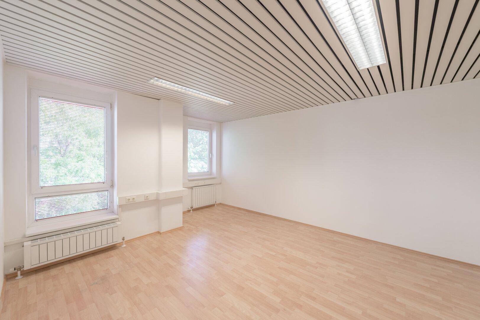 Leerer Büroraum mit hellen Wänden, Holzoptikboden und Fenstern mit Jalousien für Privatsphäre.