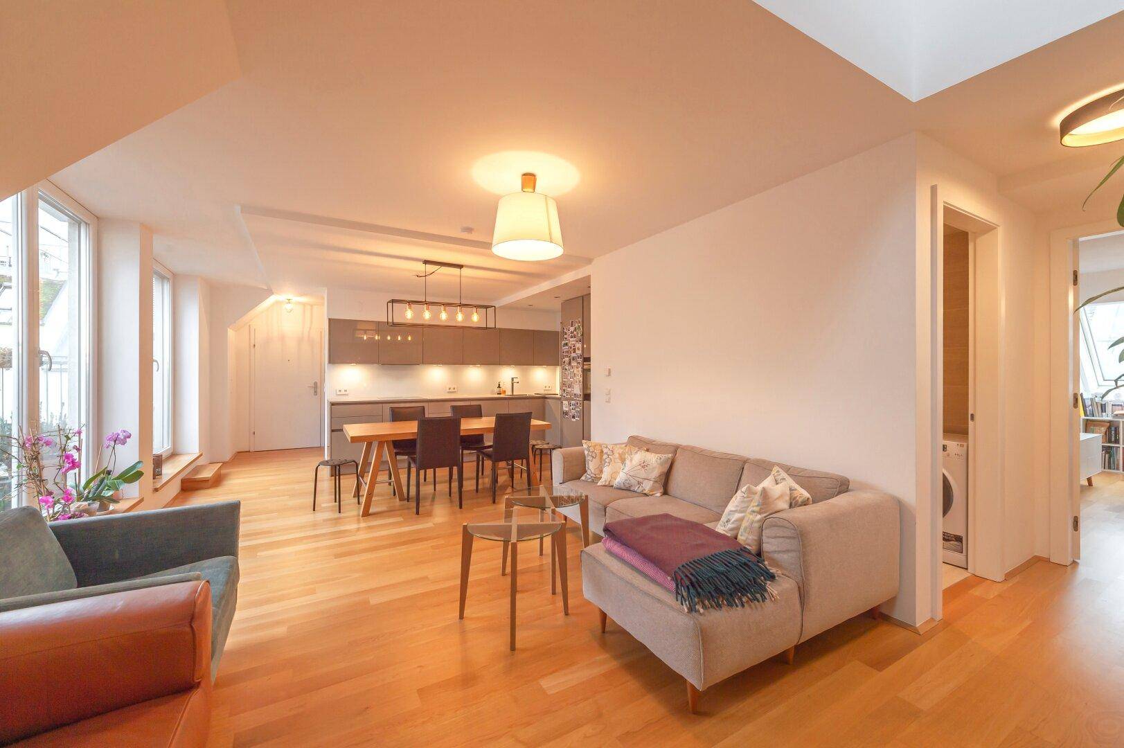 Heller Wohnbereich mit Sofa, Esstisch und offener Küche, verbunden mit einem Flur.