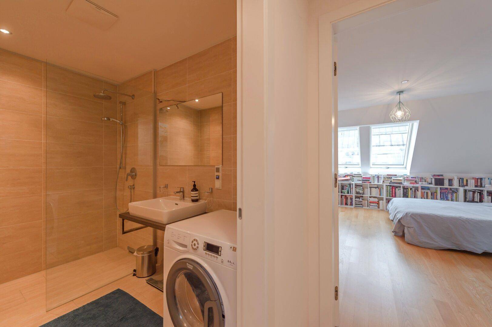 Modernes Badezimmer mit begehbarer Dusche und Waschmaschine, angrenzend an ein Schlafzimmer.