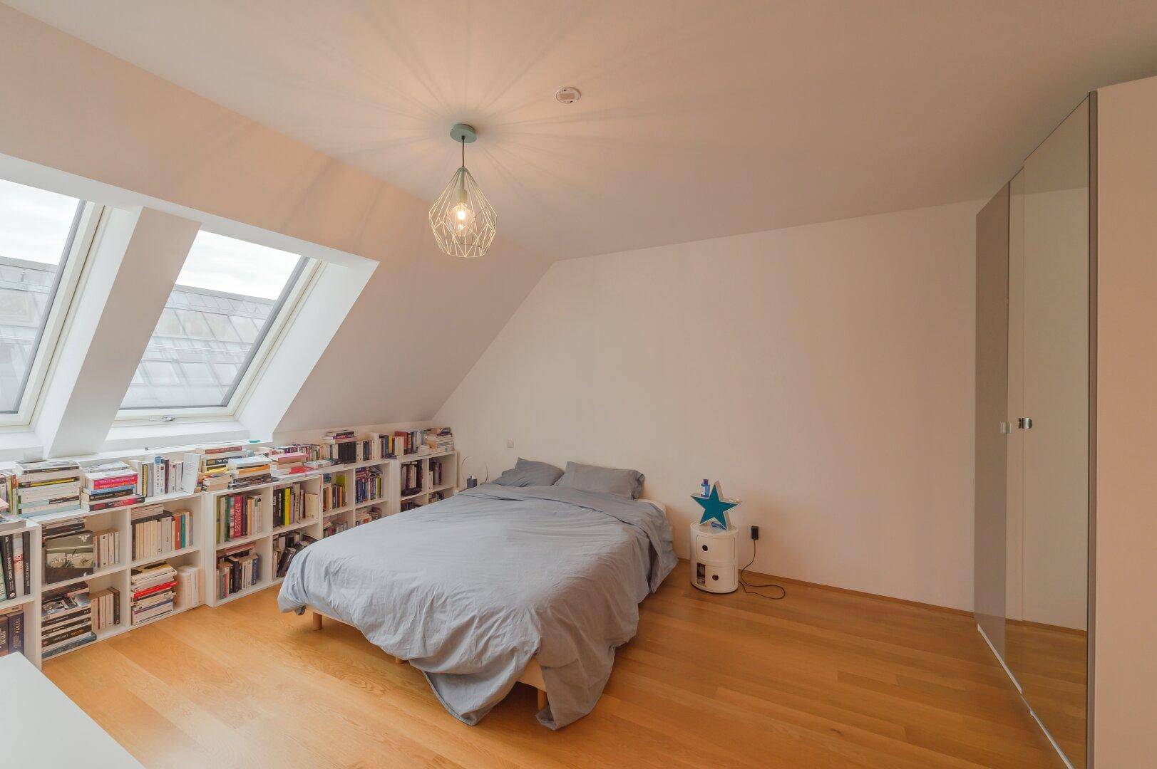 Helles Schlafzimmer im Dachgeschoss mit schrägen Wänden, Dachfenstern und Bücherregalen.