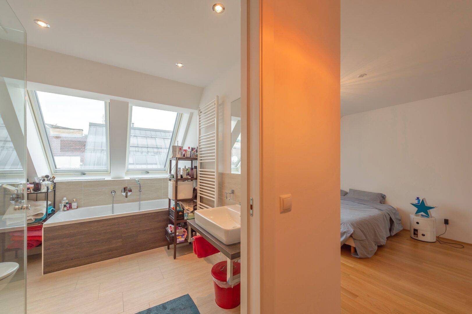 Modernes Badezimmer mit Badewanne, Waschbecken und Dachfenstern, angrenzend an ein Schlafzimmer.