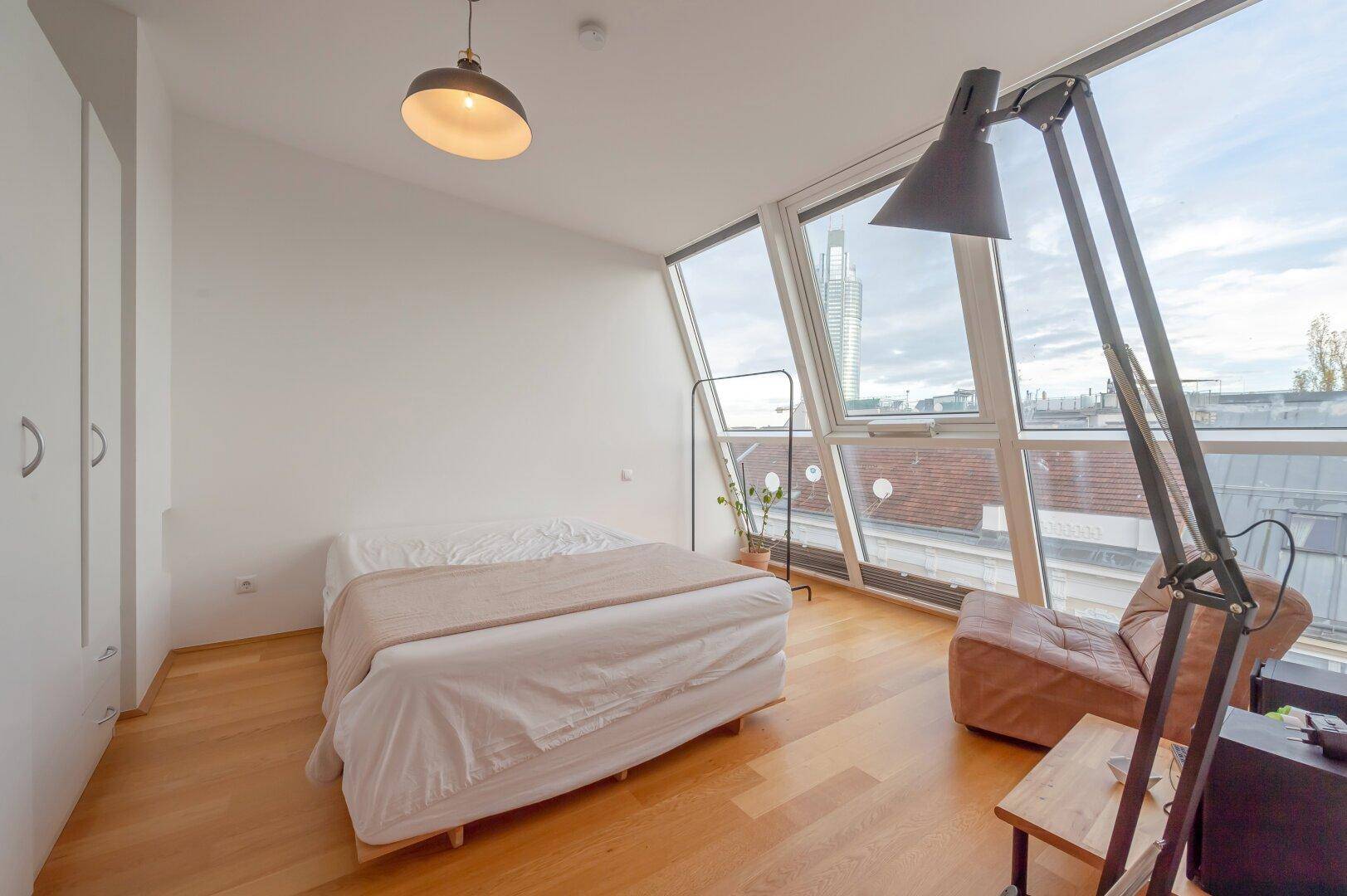 Helles Schlafzimmer mit Holzboden, großen Dachfenstern und Blick auf die Stadt.