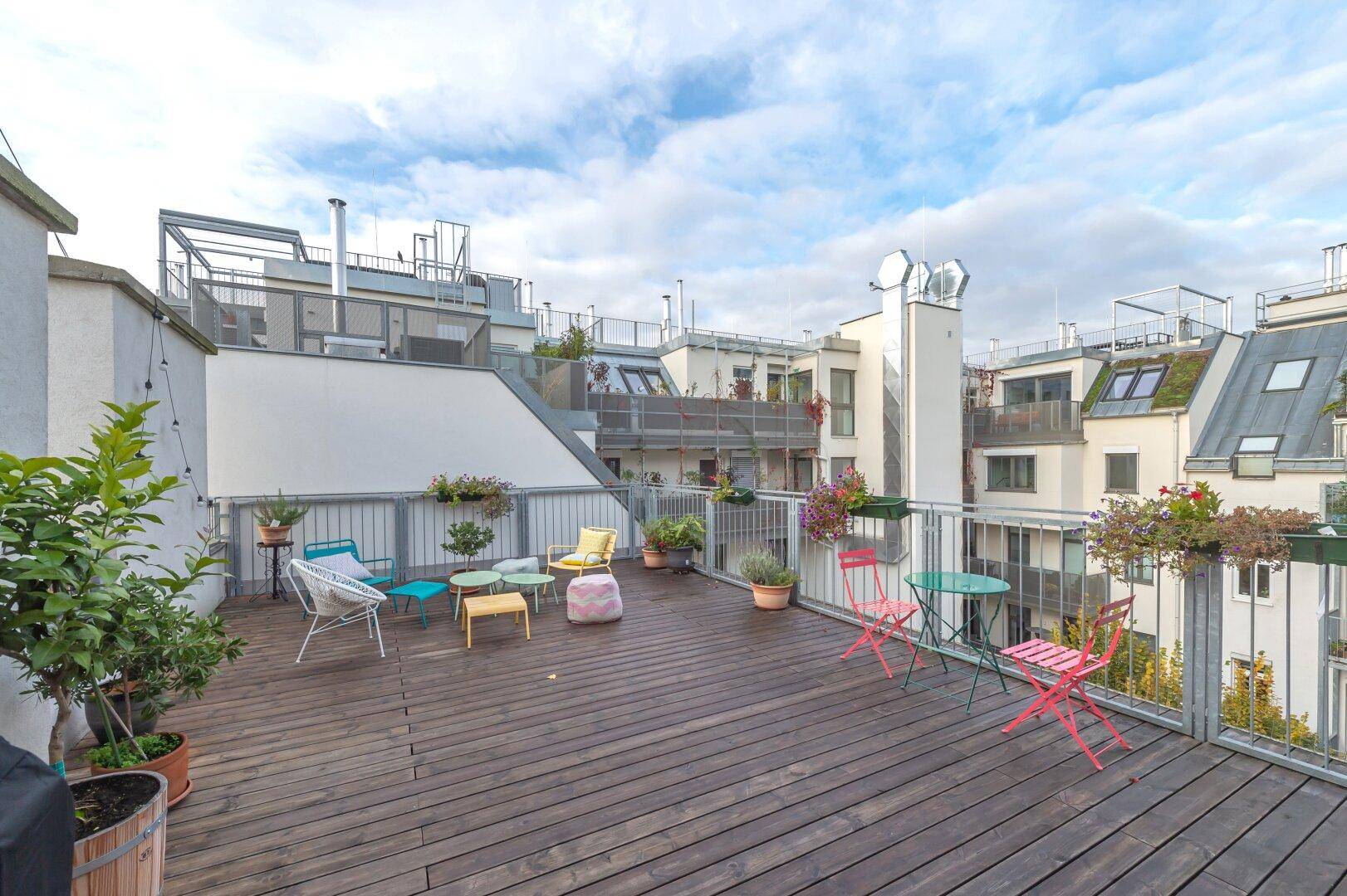 Geräumige Dachterrasse mit Holzboden, bunten Gartenmöbeln und Blick auf die umliegenden Gebäude.
