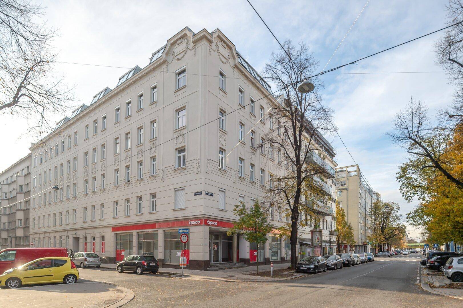 Eckgebäude an einer belebten Straße mit Geschäften im Erdgeschoss und Bäumen.