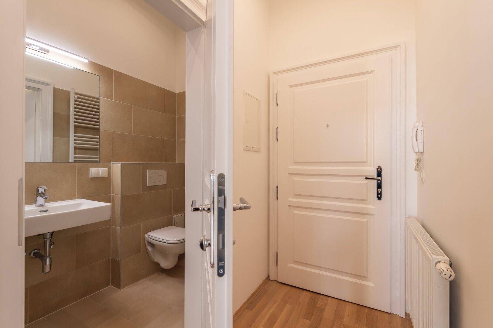 Modernes Gäste-WC mit Wand-WC, Waschbecken und beigefarbenen Fliesen, vom Flur zugänglich.