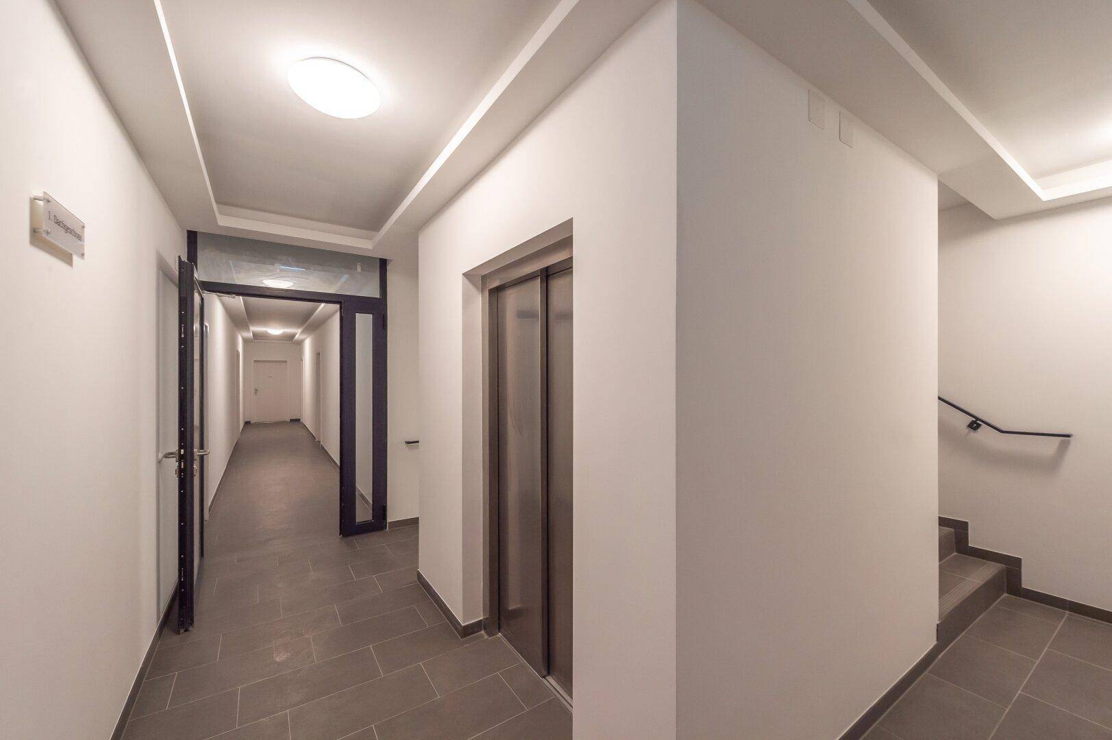 Moderner Hausflur mit Aufzug, Treppenhaus und dunklen Fliesen im Eingangsbereich.