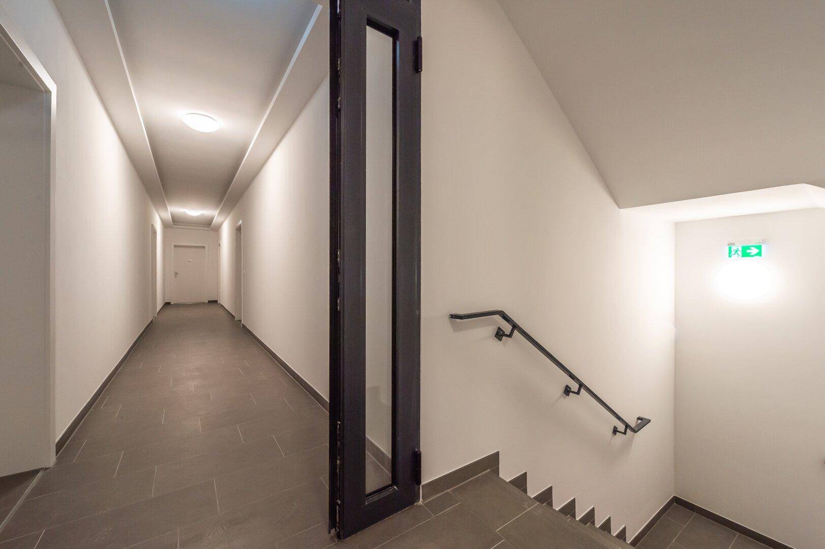 Heller Hausflur mit Aufzug und Treppenhaus, ausgestattet mit dunklen Bodenfliesen.