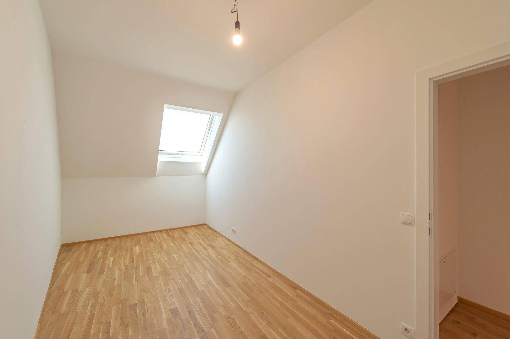 Helles Zimmer mit Dachschräge, Holzboden und einem großen Dachfenster für viel Tageslicht.