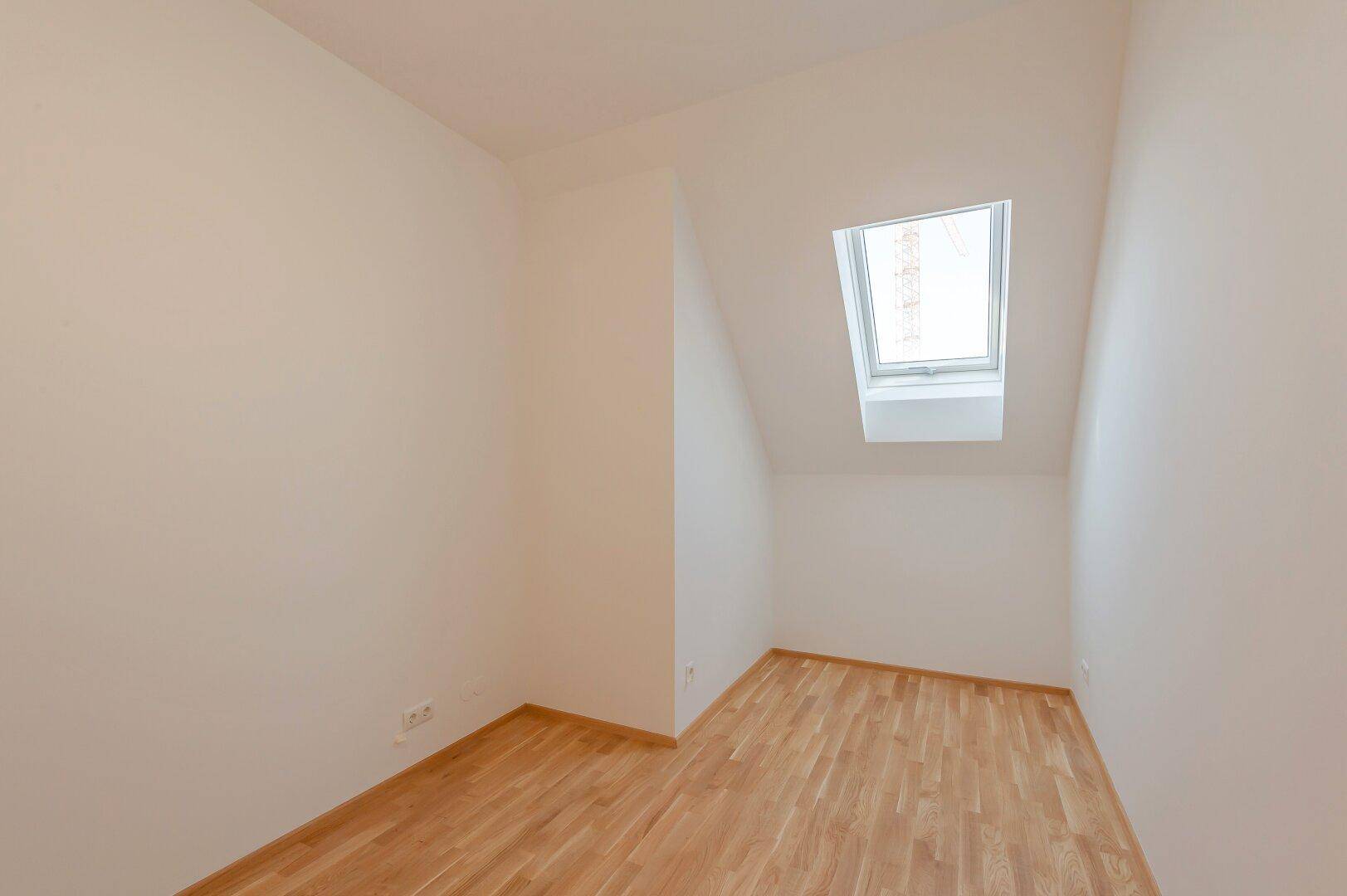 Helles Zimmer mit Dachschräge, Holzboden und einem großen Dachfenster für viel Tageslicht.