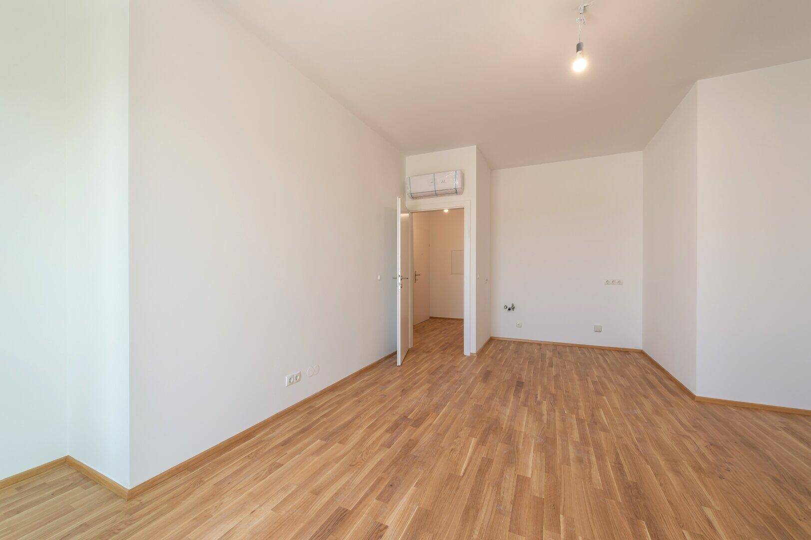 Helles Zimmer mit Holzboden, weißen Wänden und einer Klimaanlage über der Tür.