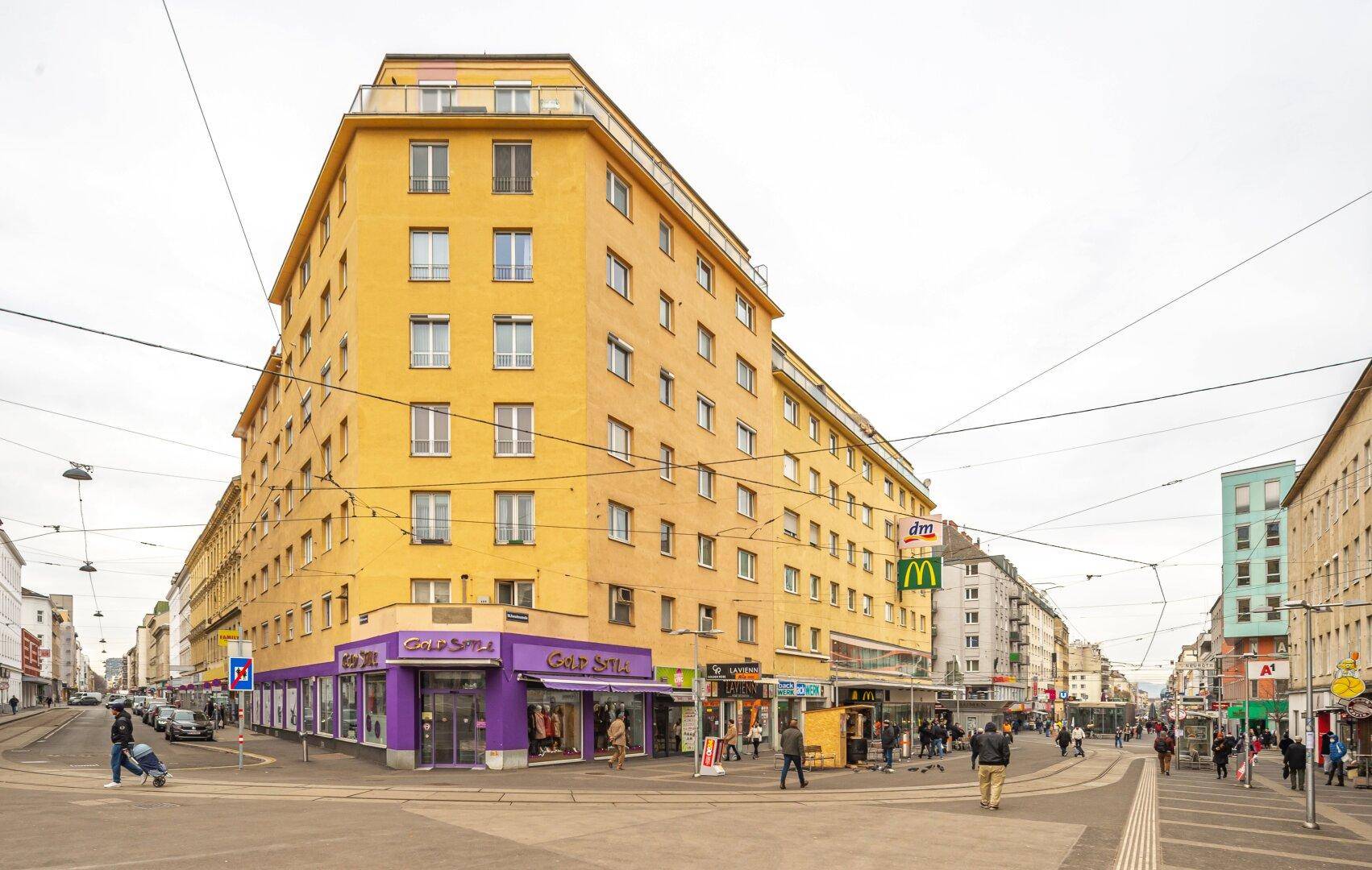 Gelbes Mehrfamilienhaus an einer belebten Straßenecke mit Geschäften im Erdgeschoss.