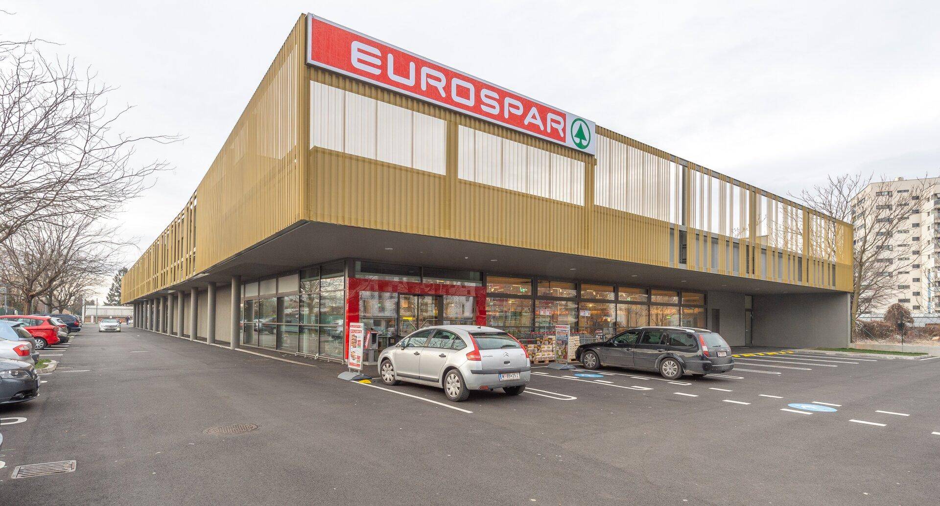 Eingangsbereich des Eurospar-Supermarktes mit geparkten Autos und großen Schaufenstern.