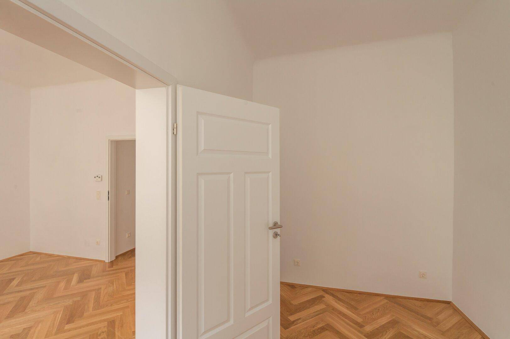 Heller Flur mit Fischgrätparkett und weißer Tür, der zu weiteren Räumen führt.