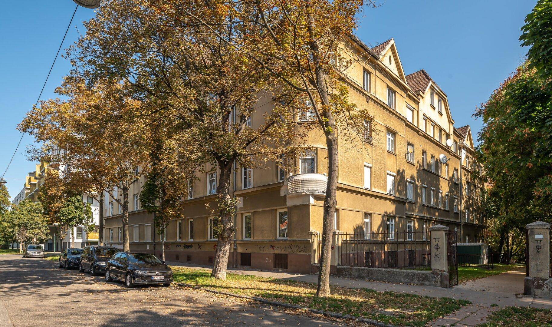 Wohngebäude mit Bäumen und parkenden Autos an der Straße unter blauem Himmel.