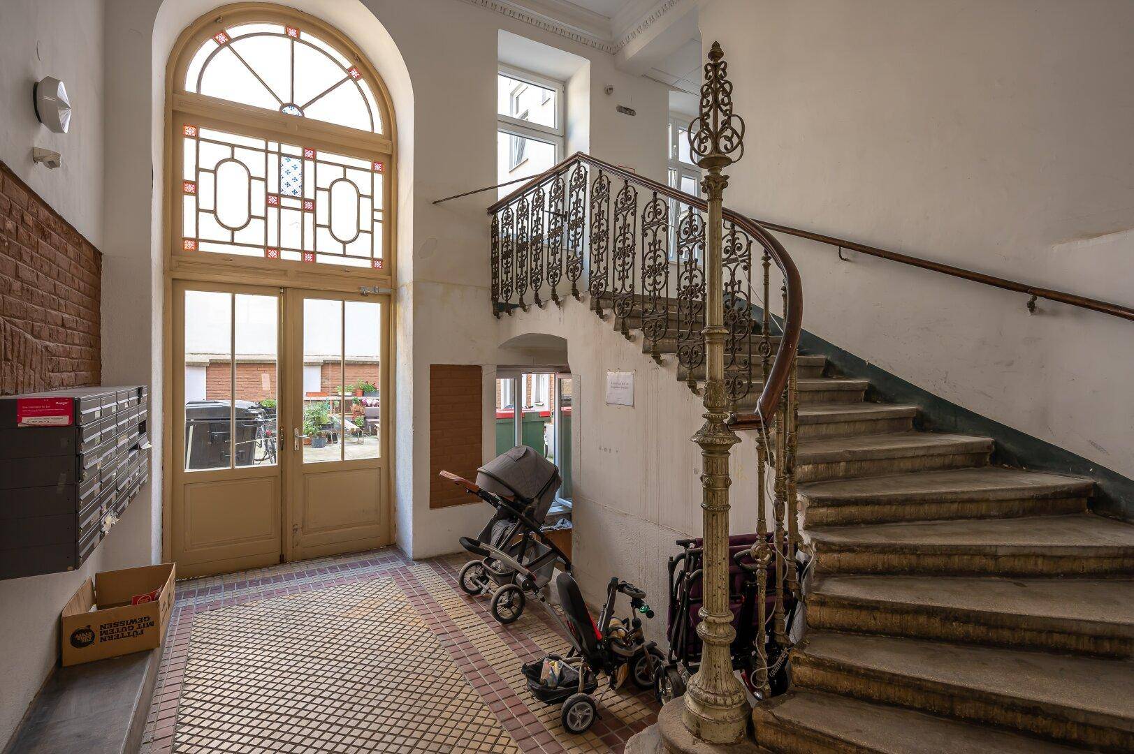 Historisches Treppenhaus mit schmiedeeisernem Geländer und originalen Fliesen im Eingangsbereich.