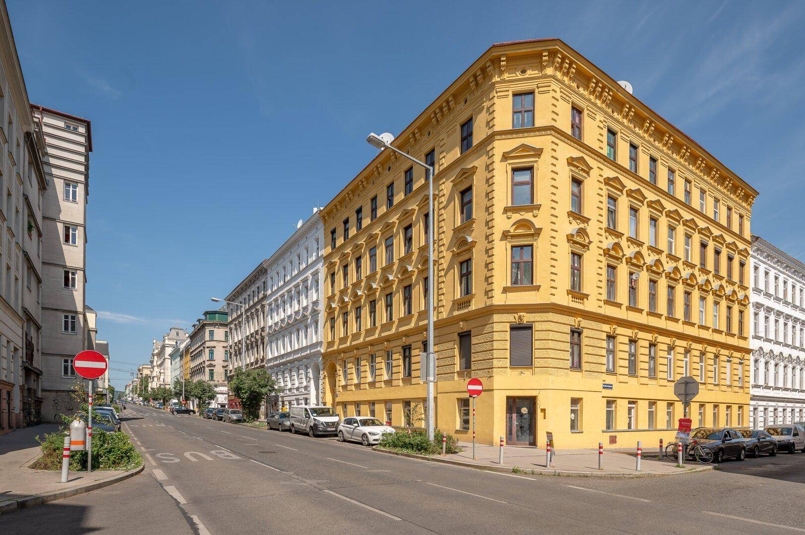 Historisches Eckgebäude mit gelber Fassade und vielen Fenstern an einer belebten Stadtstraße.