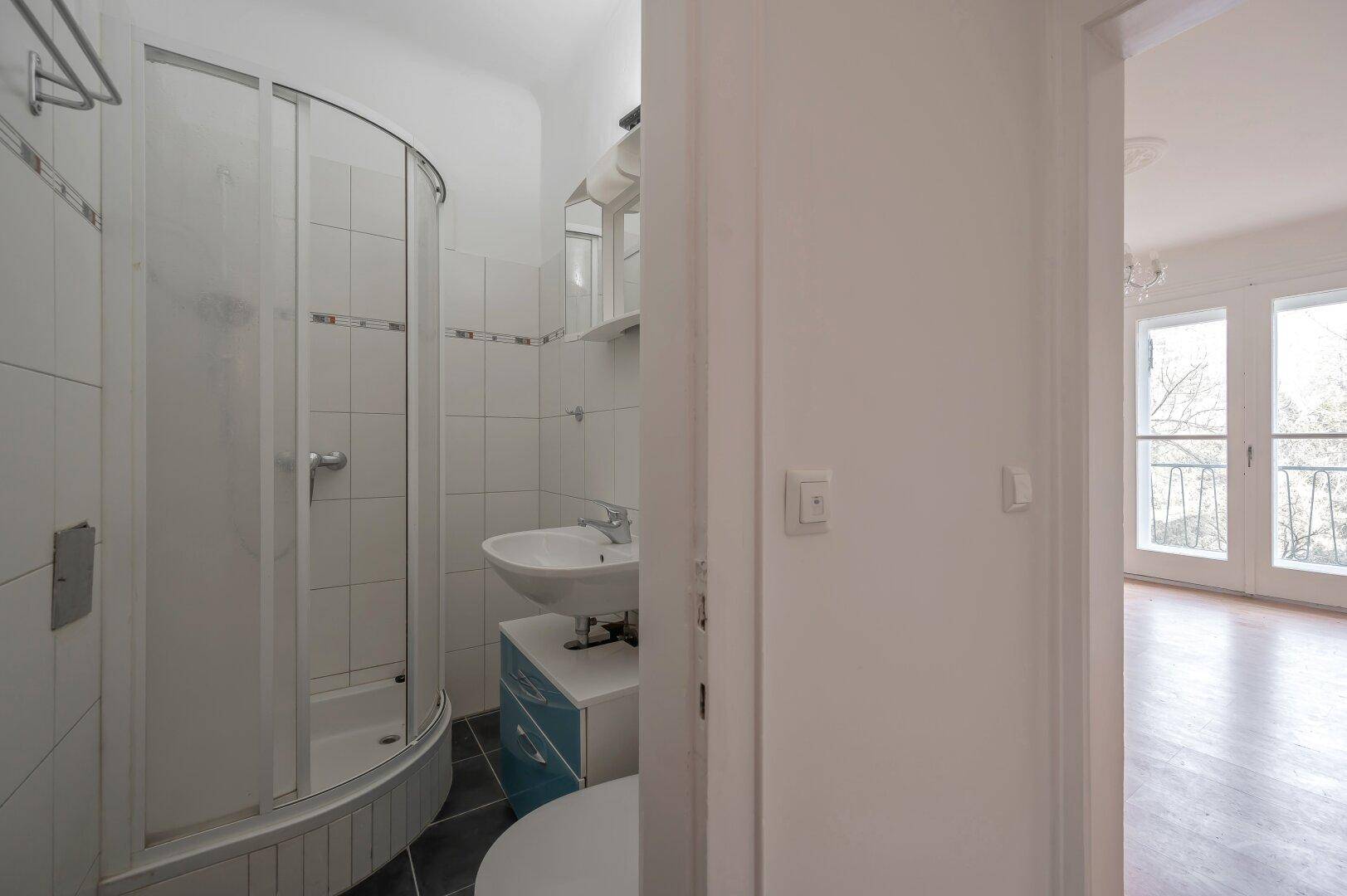 Badezimmer mit Duschkabine und Waschbecken, vom Flur aus sichtbar.
