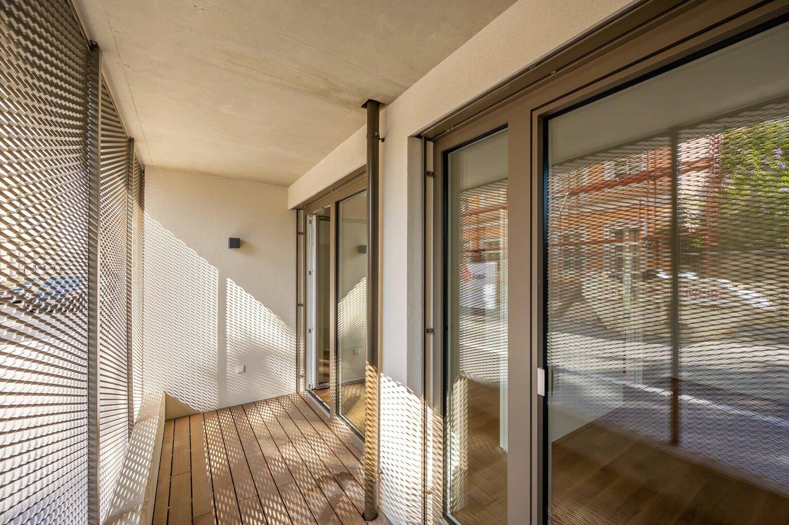 Sonniger Balkon mit Holzboden und moderner Metallgitter-Brüstung, bietet einen Blick nach draußen.