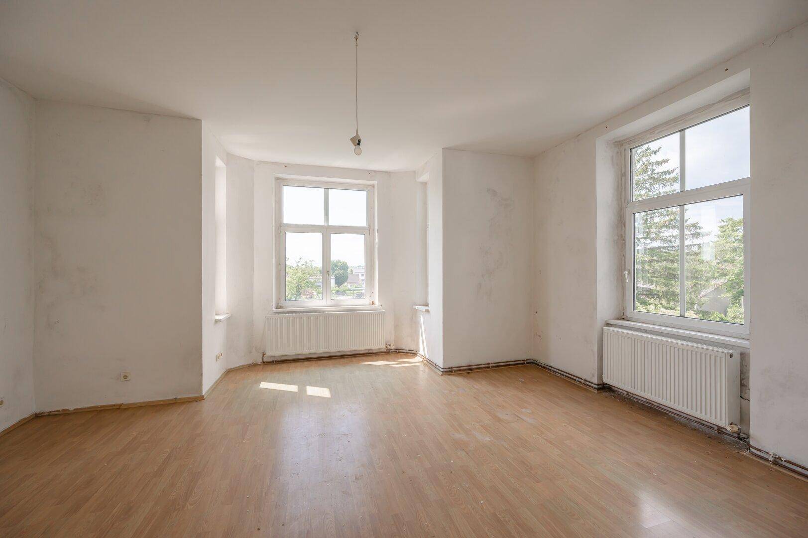 Helles, leeres Zimmer mit Laminatboden und zwei großen Fenstern, bereit zur Renovierung.