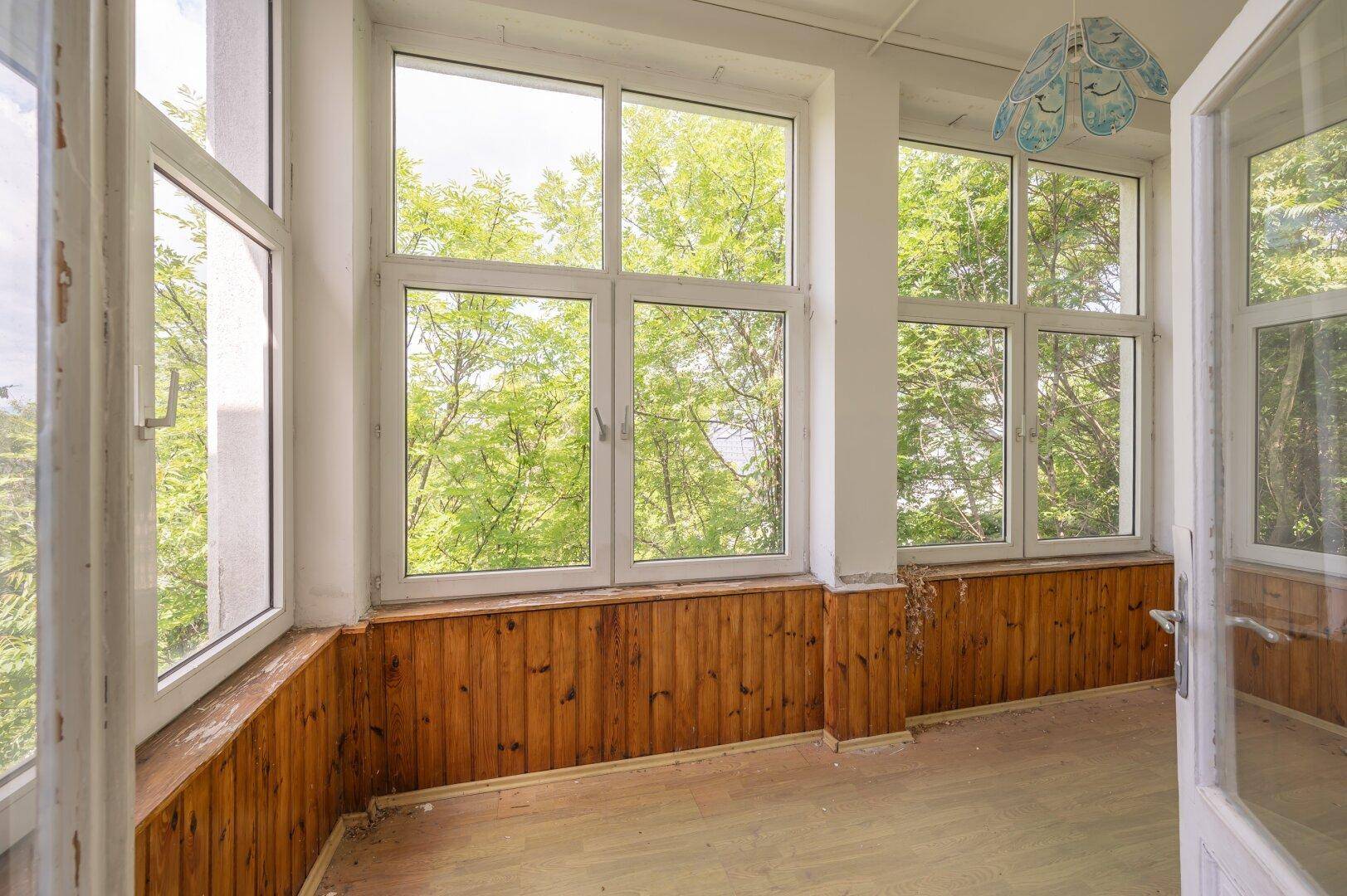 Verglaster Wintergarten mit Holzvertäfelung und weitem Blick ins Grüne, viel Tageslicht.