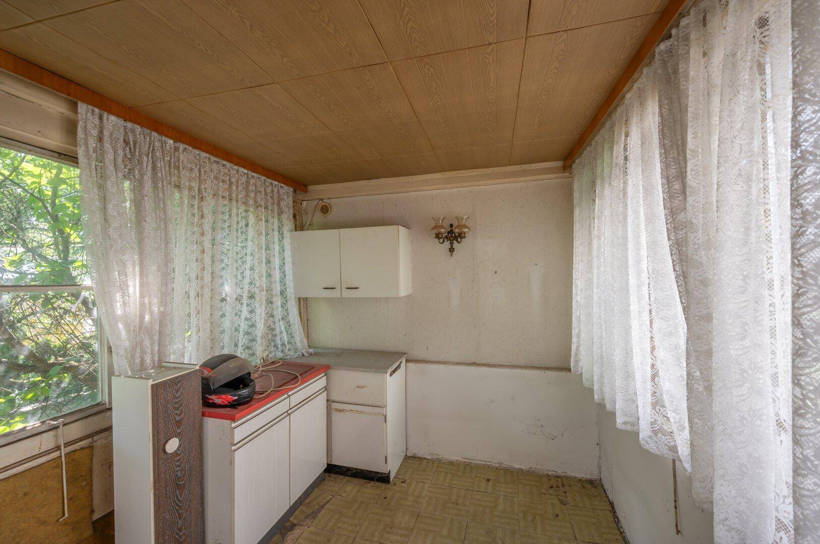 Kleine Küchenzeile mit Vorhängen und Holzdecke, die eine umfassende Modernisierung benötigt.