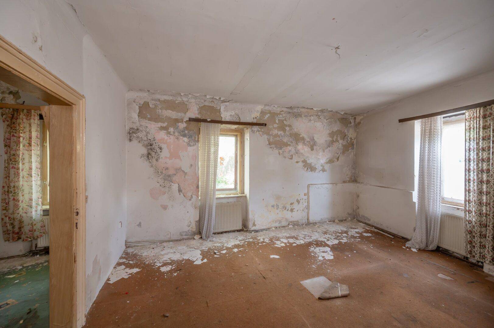 Schlafzimmer mit abblätternder Tapete und zwei Fenstern, das eine vollständige Renovierung benötigt.