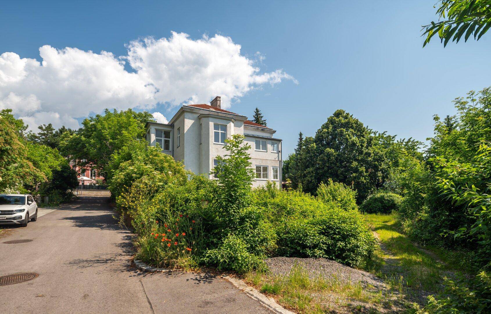 Historisches Gebäude an einer Straße, umgeben von Bäumen und Sträuchern unter blauem Himmel.
