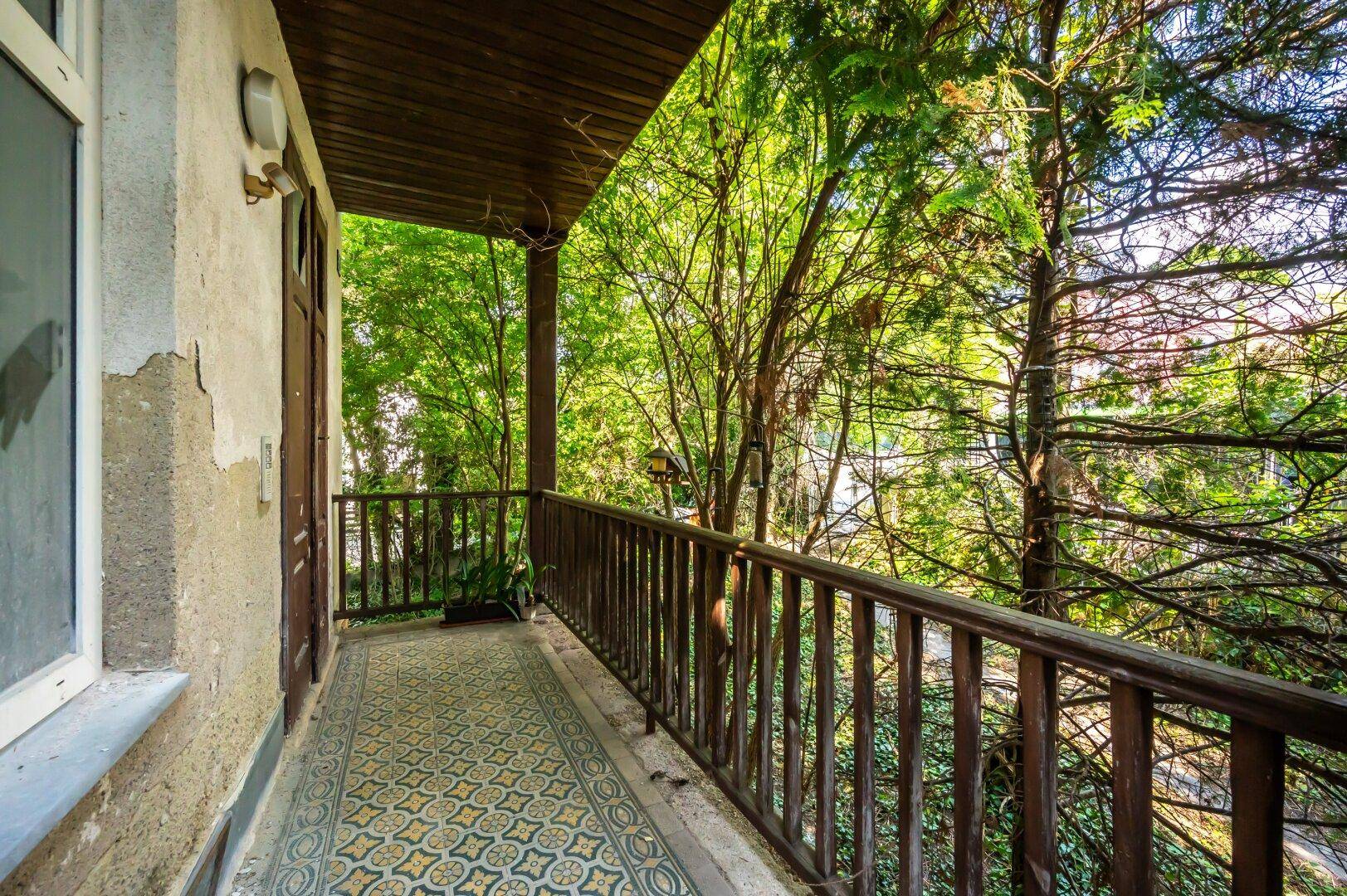 Überdachter Balkon mit gemusterten Fliesen und Holzbalken, umgeben von dichtem Grün.