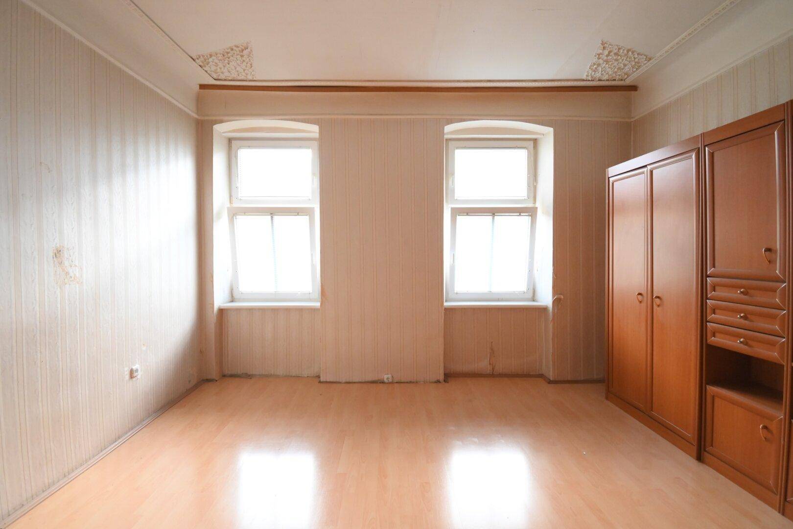 Geräumiges Zimmer mit Laminatboden, zwei Fenstern und einem großen Holzschrank an der Wand.