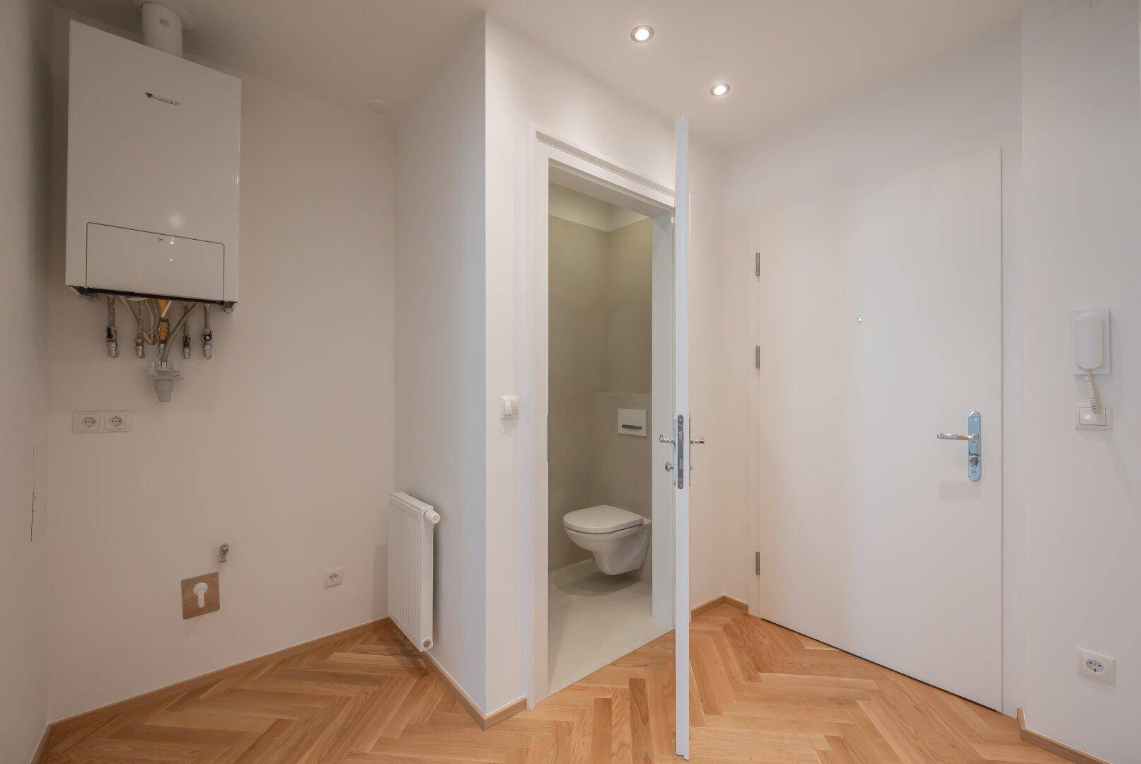 Heller Eingangsbereich mit Fischgrätparkett, Heiztherme und Zugang zu einem Gäste-WC.
