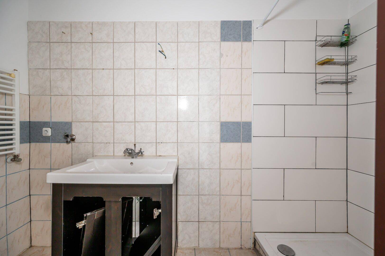 Badezimmer mit gefliesten Wänden, Waschbecken und Duschbereich, in renovierungsbedürftigem Zustand.