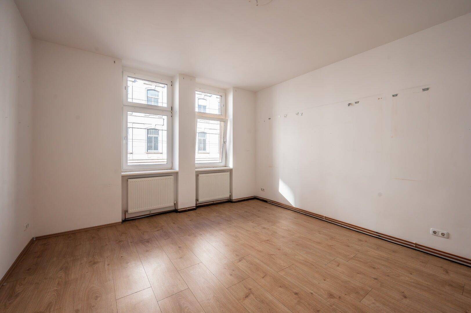 Leeres Zimmer mit Holzboden und zwei Fenstern, bereit zur individuellen Gestaltung.