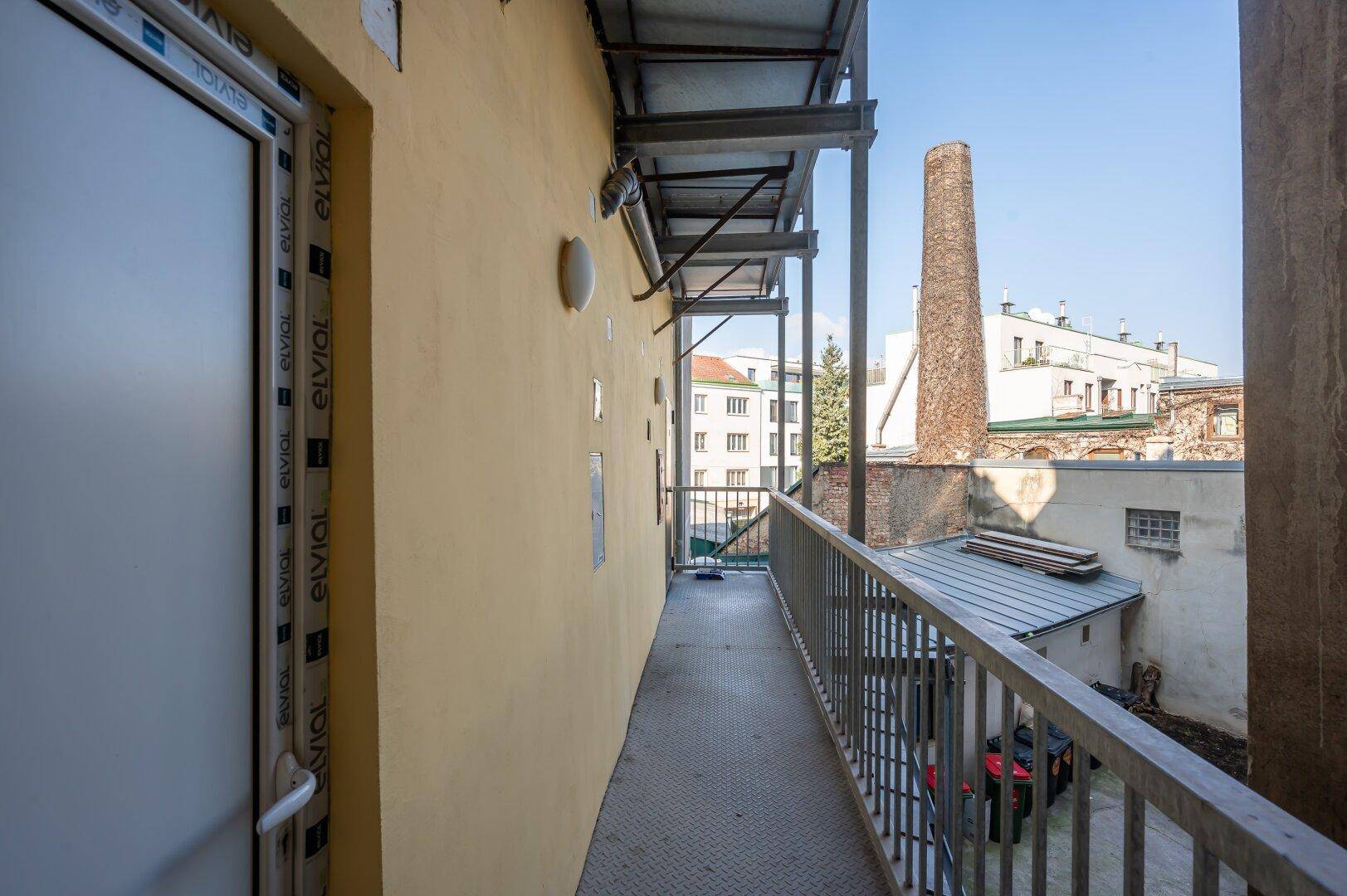 Langer Außenbalkon mit Metallgeländer und Blick auf städtische Gebäude und einen Schornstein.
