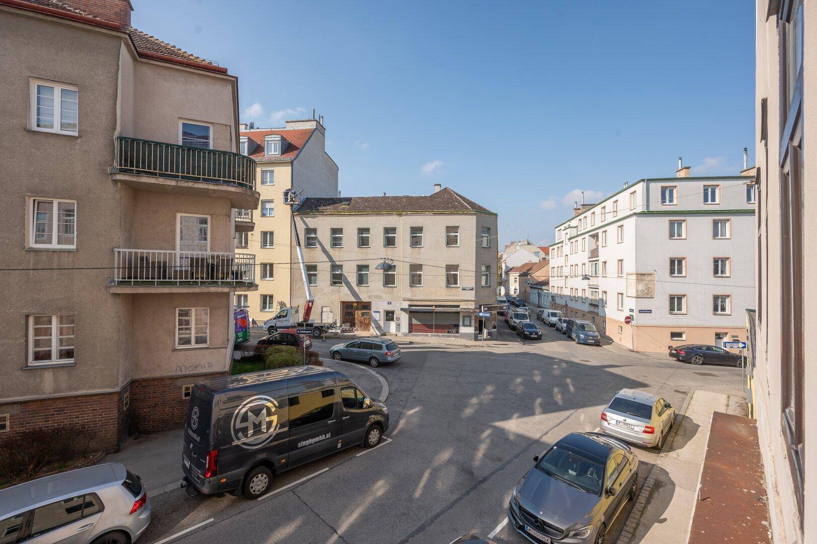 Belebte Stadtstraße mit Gebäuden, geparkten Fahrzeugen und einem Umzugswagen an einem sonnigen Tag.